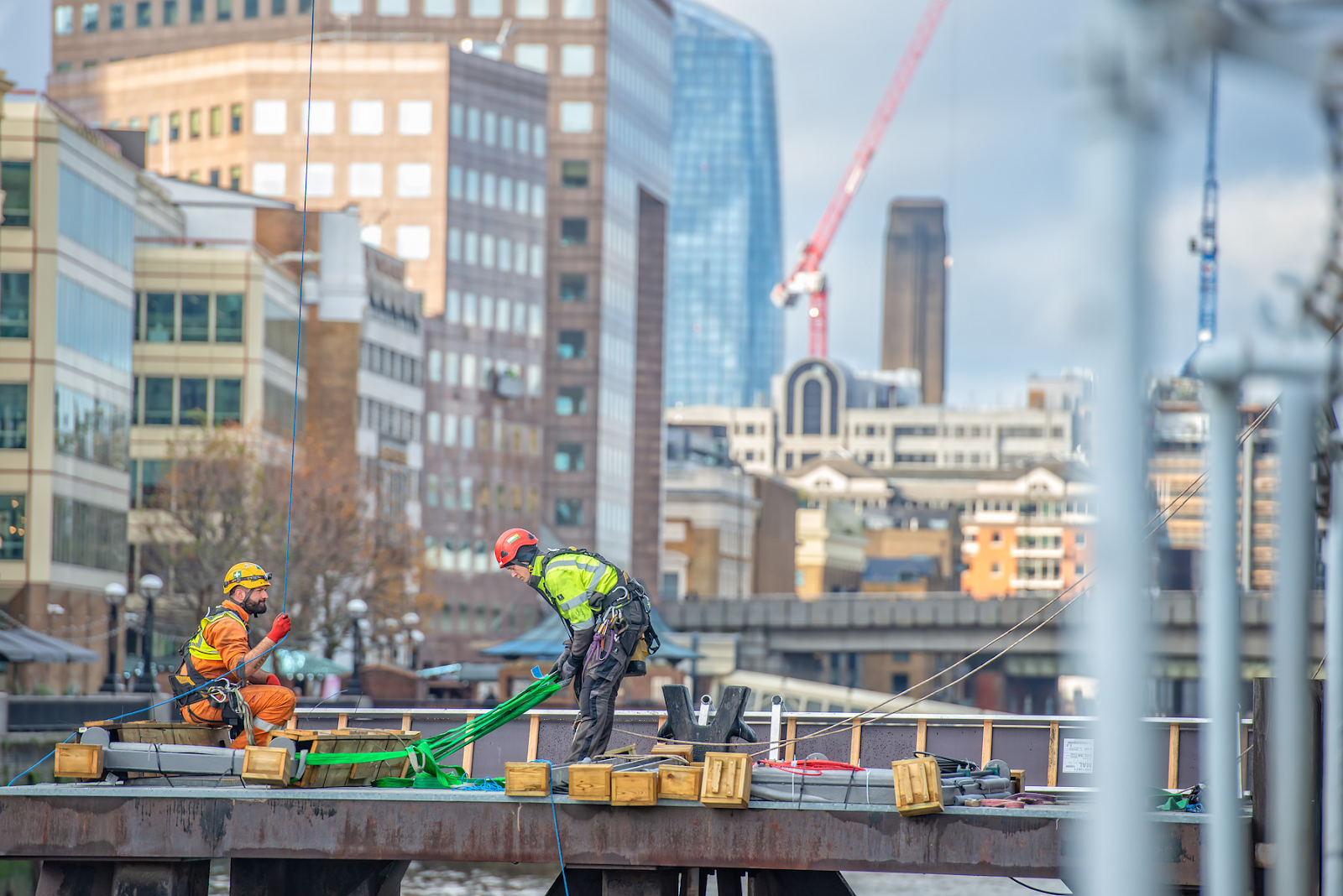 Two rope access technicians in safety gear working on a metal platform by the river with city skyscrapers in the background.