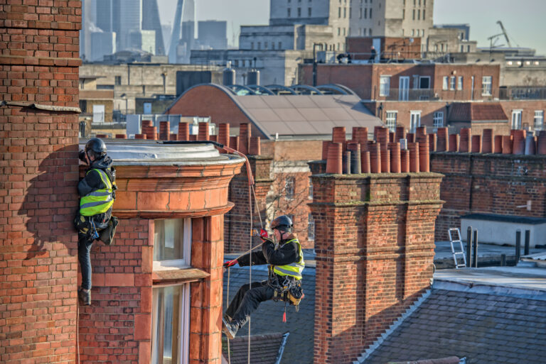 Two rope access workers repairing the facade of a red brick building in London using safety harnesses.