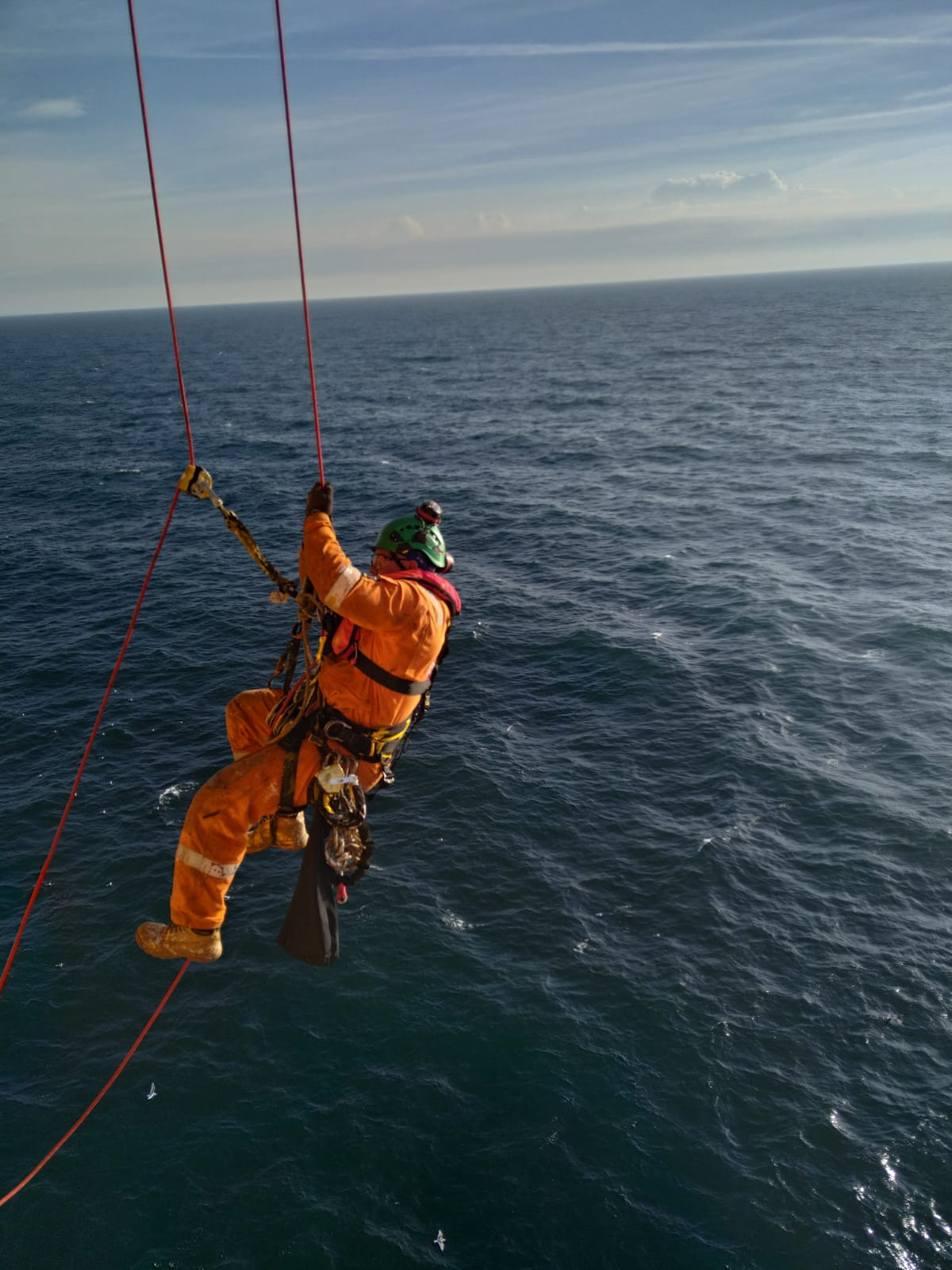 Rope access worker in orange safety gear performing maintenance over open ocean using suspended ropes.