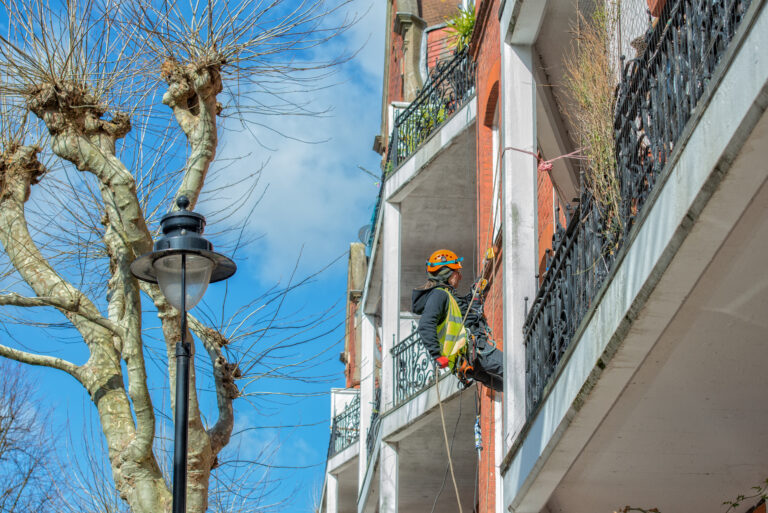 Worker using rope access to repair exterior walls of a residential building next to leafless trees.