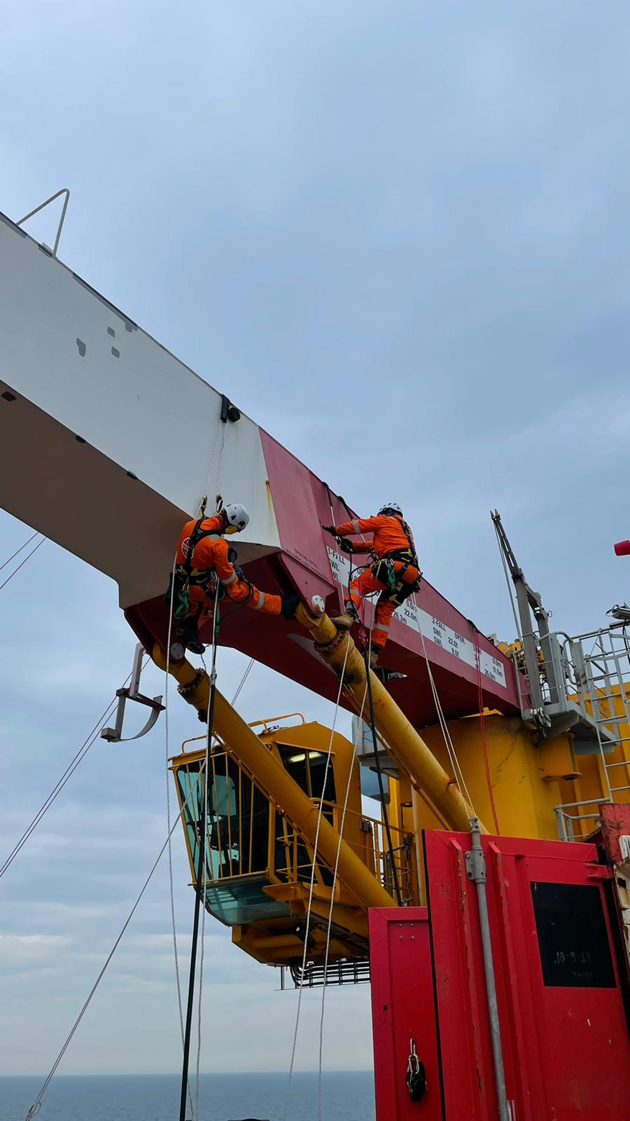 Two workers in orange suits performing rope access maintenance on a yellow and red offshore crane above the ocean.