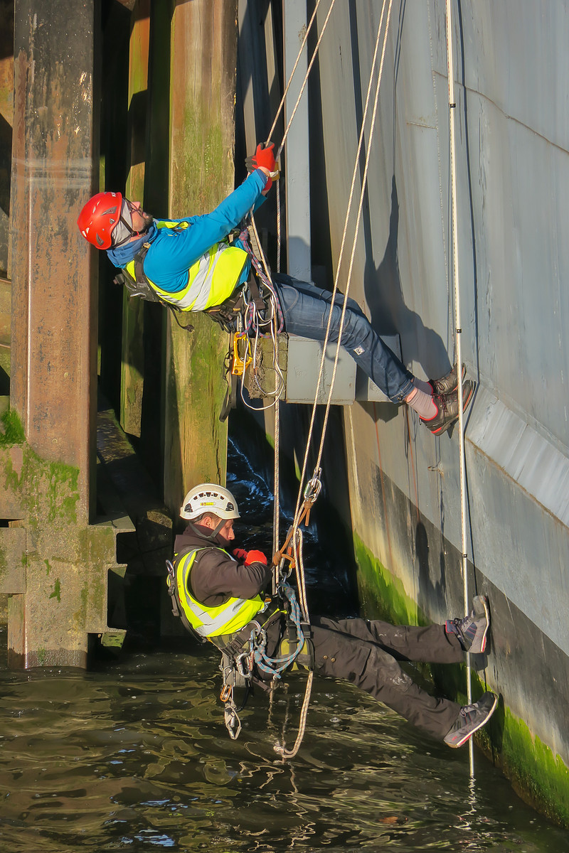 Two workers in safety harnesses suspended by ropes inspecting a metal structure above the water.