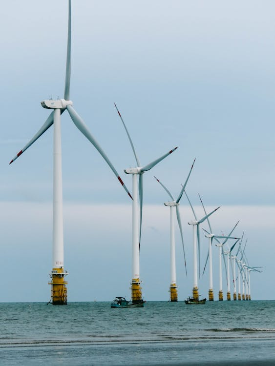 Row of wind turbines in the sea with boats nearby under a cloudy sky.