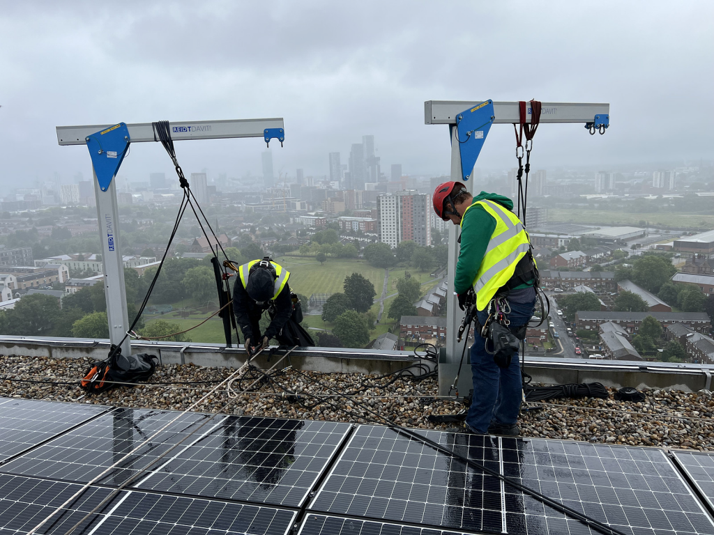 Two workers in safety gear performing maintenance on solar panels on a rooftop, with a city skyline in the background.