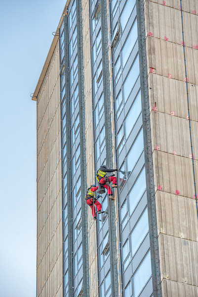 Two window cleaners in red safety gear rappelling down the side of a high-rise building, cleaning the glass windows.